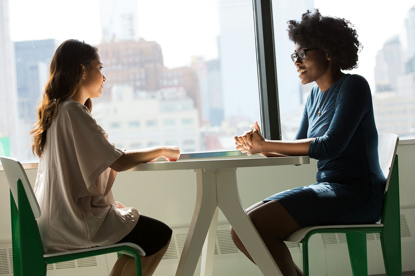 Twee vrouwen in overleg zitten aan een ronde tafel voor een groot raam.